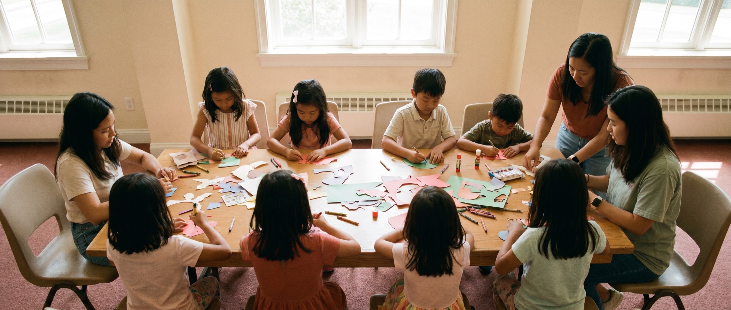 Children seated around a long table making crafts together, with two leaders helping.