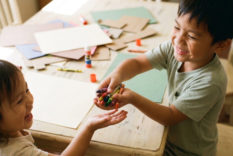 A boy handing a fistful of crayons to a smaller child across a craft table.